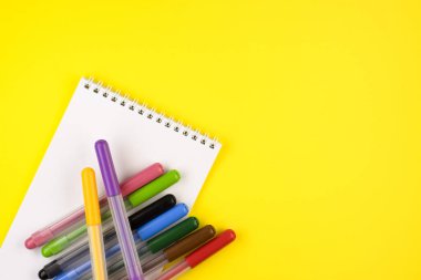 Sarı arkaplan üzerinde beyaz bir not defteri olan farklı renk işaretleri, üst görünüm. Defteri olan renkli kalemler. Kapatın.