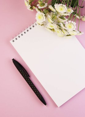 Boş bir Not Defteri, bir kalem ve pastel pembe arka planda bir çiçek buketi. Mor arka planda papatyalar olan beyaz defter.