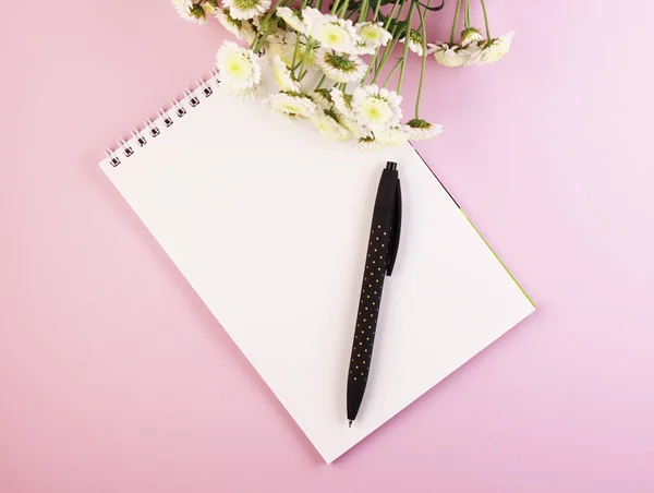 an empty Notepad with a pen and a flower bouquet on a pastel pink background. white notebook with daisies on a purple background