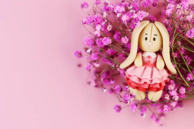 Pembe arka planda plastik Paskalya Tavşanı. Güzel bir elbise içinde, bir buket mor çiçeğin yanında sevimli bir tavşan.