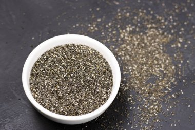 Siyah taşlı arka planda beyaz bir kasede siyah Chia tohumları. Küçük İspanyol adaçayı taneleri, fasulyeye benzer, tadı fındık gibi, gri-beyaz-siyah renkli ve kabartma desenli. sağlık ürünü