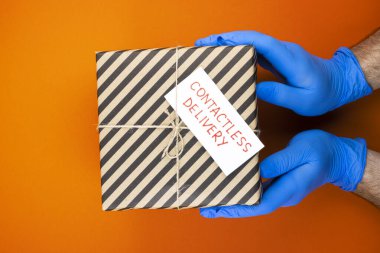 Paketlerin teslimatı yok. Tıbbi eldivenli eller, paketi turuncu arka planda tutuyor. Kapıya teslimat. Karantina sırasında yiyecek teslimatı