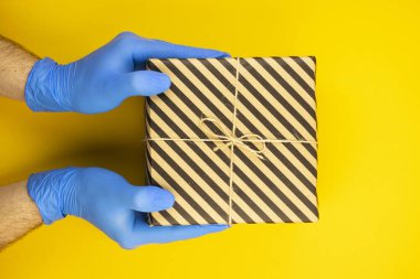 Temassız teslimat kavramı. Sarı arka planda eldivenlerin elinde bir paket var. Paketleri güvenli bir şekilde kapıya teslim et.