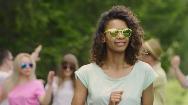 Flirty Girl mit lockigen Haaren tanzen, lächelnd in die Kamera. Freunde feiern im Park — Stockvideo