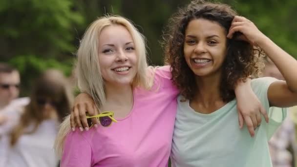 Две привлекательные девушки обнимаются, смотрят в камеру, молодые люди веселятся — стоковое видео