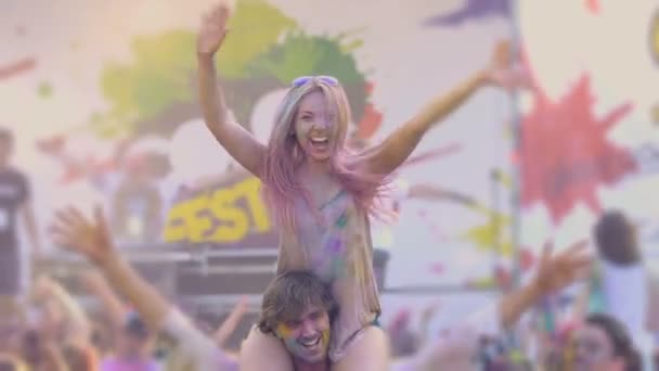 Beautiful woman sitting on boyfriend's shoulders, waving hands at Holi festival — Stock Video