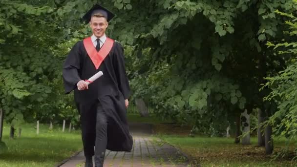 Feliz graduado sonriente caminando en el parque con diploma, saltando con emoción — Vídeos de Stock