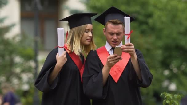 Amigos graduados con vestidos académicos ver fotos en el teléfono inteligente — Vídeos de Stock