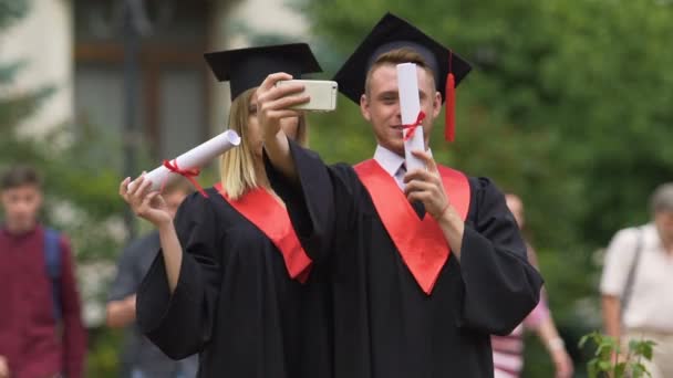 Homem e mulher em vestidos acadêmicos tirando selfie após a cerimônia de formatura — Vídeo de Stock