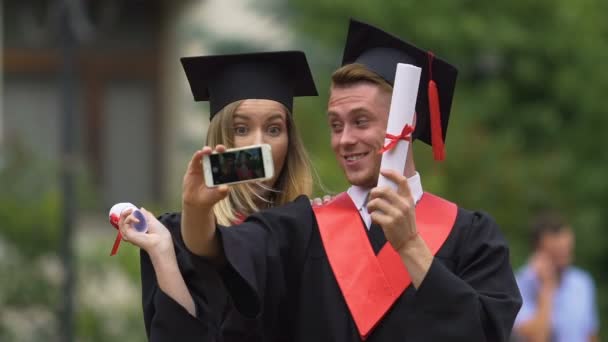 Gelukkig man en vrouw in academische caps en toga's filmen video op smartphone — Stockvideo