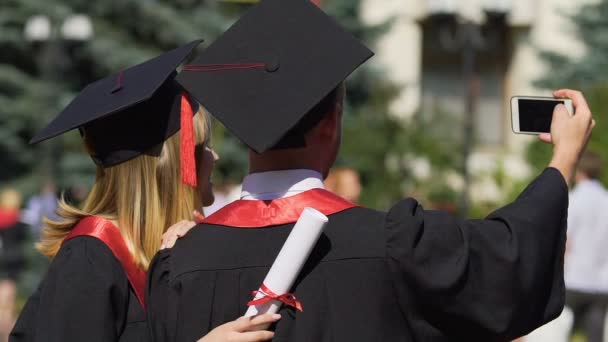 Hombre y mujer con batas y gorras académicas tomando selfie en la ceremonia de graduación — Vídeos de Stock