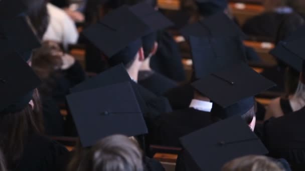 Jóvenes con sombreros académicos sentados en la sala de conferencias, ceremonia de entrega de diplomas — Vídeos de Stock