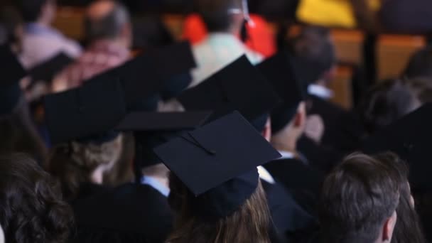 Students sitting at graduation ceremony, light balloon tied to man's mortarboard — Stock Video