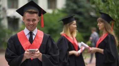 Akıllı telefon tutarak ve kameraya, mezuniyet seyir gülümseyen erkek mezunu
