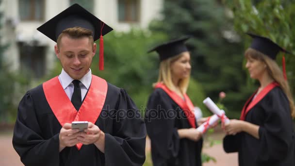 Joyous male graduate typing message on smartphone, browsing mobile applications — Stock Video