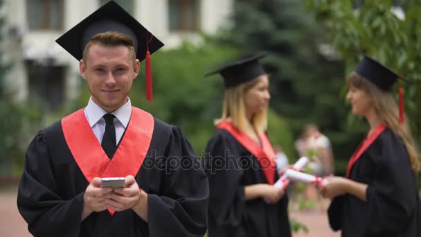 Afgestudeerde studenten staan in park, jongeman met smartphone camera kijken — Stockvideo