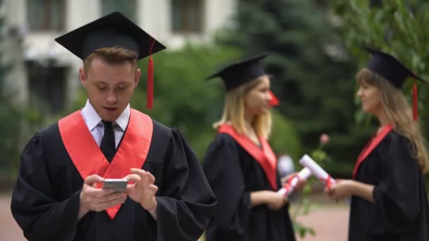 Día de la graduación, joven navegando en el teléfono inteligente y sonriendo para la cámara — Vídeo de stock