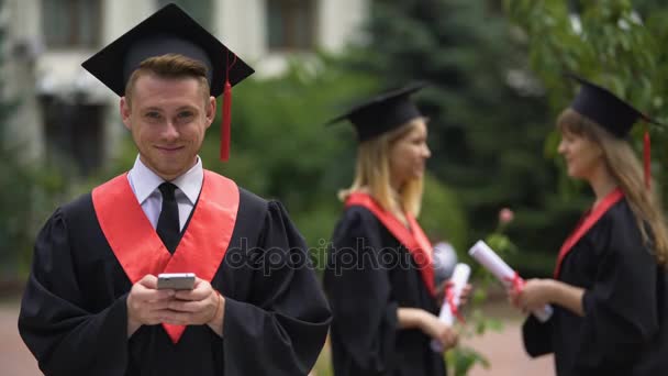 Jonge mannelijke afgestudeerde holding smartphone en lachend voor camera, afstuderen gebeurtenis — Stockvideo