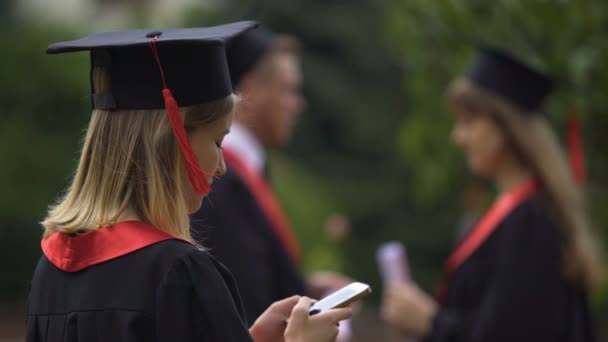 Χαμογελώντας ξανθιά απόφοιτος στέκεται κοντά φίλους και γραπτών μηνυμάτων σε smartphone — Αρχείο Βίντεο