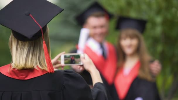 Mujer en vestido académico y sombrero filmando mejores amigos en el teléfono, día de la graduación — Vídeos de Stock