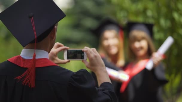 Dos chicas graduadas sosteniendo diplomas y posando para cámara de teléfono inteligente, graduación — Vídeo de stock