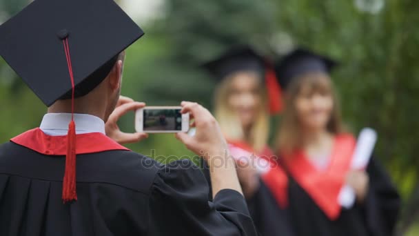 Молодой выпускник в мантии и кепке фотографирует девушек по мобильному телефону, дружит — стоковое видео