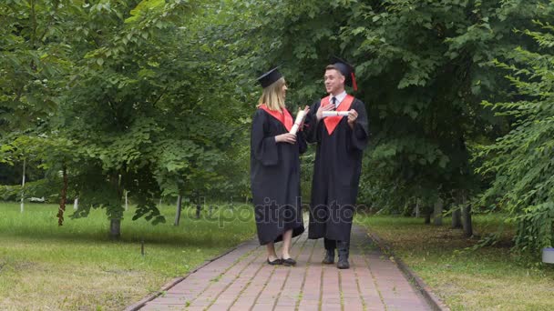 Junge glückliche Absolventen, die Diplome halten und im Park spazieren gehen, reden und entspannen — Stockvideo