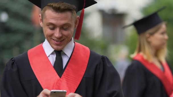 Neşeli erkek lisansüstü akademik elbiseli Smartphone'da sohbet ve gülüyor — Stok video