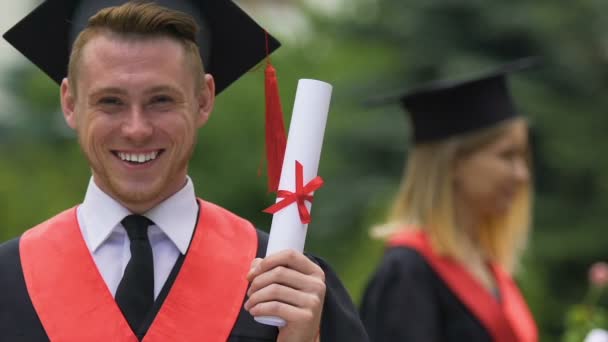 Male graduate in academic dress holding university diploma, smiling to camera — Stock Video