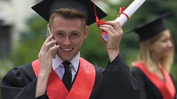 Glimlachend mannelijke afstuderen student holding universitair diploma, praten over de telefoon — Stockvideo