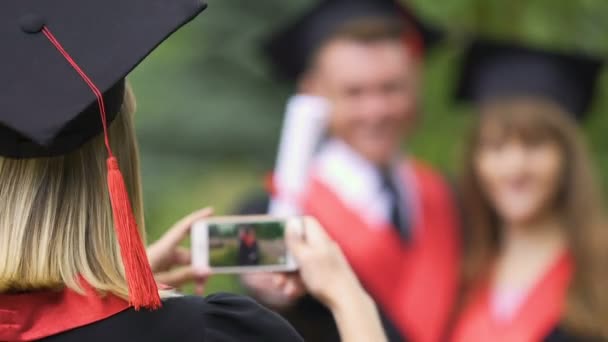 Jovencita en vestido académico tomando fotos de amigos graduados de la universidad — Vídeo de stock