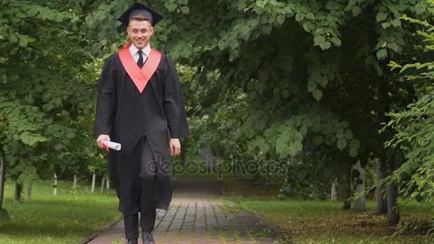 Усміхнений молодий чоловік в академічній сукні, що йде і стрибає, шлях до майбутнього успіху — стокове відео