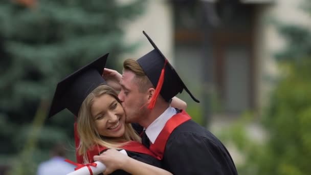 Lento-mo de casal em vestido acadêmico beijos na bochecha, jogando com certificados — Vídeo de Stock