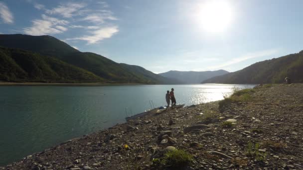 Пара прогуливающихся вдоль берега солнечной реки, держащихся за руки, горы на горизонте — стоковое видео