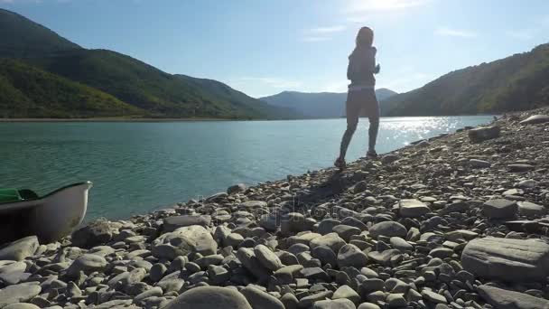 晴れた日に山の小石の太陽に照らされた川岸に沿ってジョギングする若い女性 — ストック動画