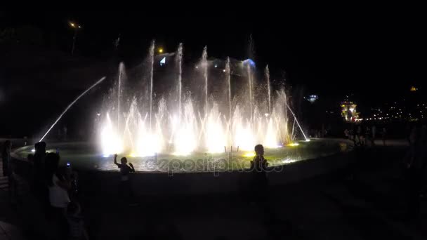 Silhouetten van mensen in de buurt van verlichte fontein, water spatten in de lucht — Stockvideo