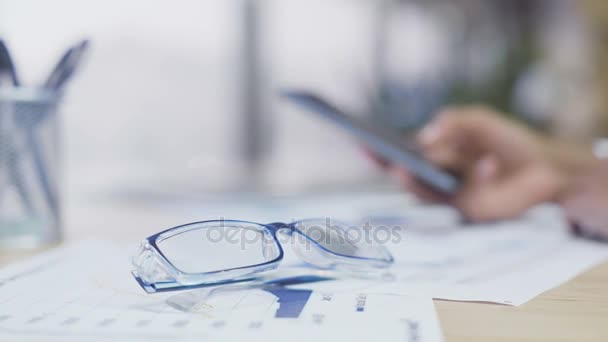 Eyeglasses lying on the table, woman scrolling pages on smartphone, office break — Stock Video