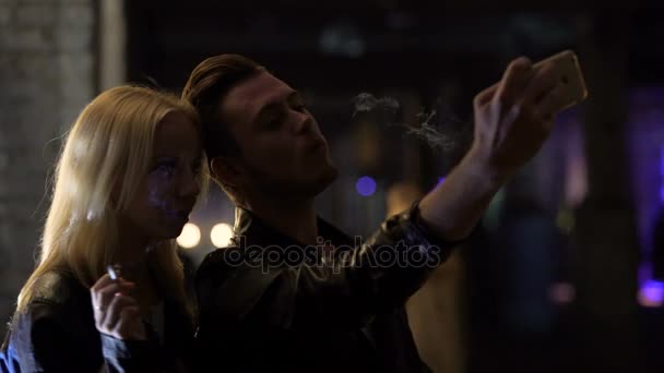 Молодой человек и женщина курят и делают селфи на смартфоне в ночном клубе — стоковое видео
