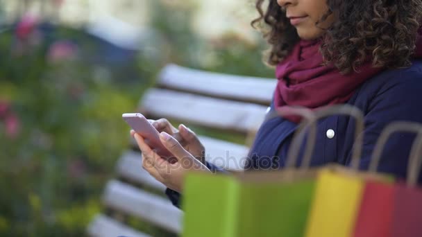 Joven dama de compras en las tiendas en línea en el teléfono inteligente, sentado en el banco al aire libre — Vídeos de Stock