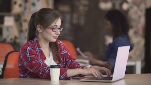 Жінка-журналістка в окулярах сидить в кафе і друкує на ноутбуці, фрілансер — стокове відео