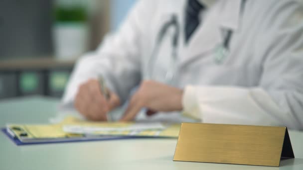 Médico atento prescribiendo medicamentos en la clínica, plantilla de placa de identificación en la mesa — Vídeo de stock