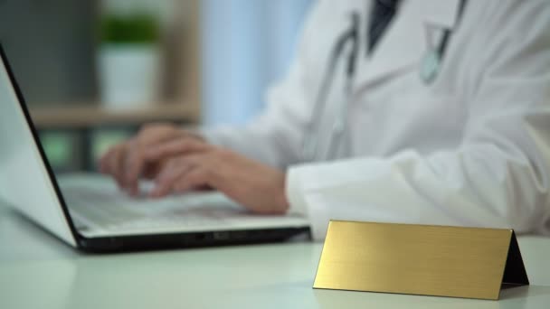 Terapeuta en uniforme escribiendo información médica en el ordenador portátil, placa de identificación en blanco en la mesa — Vídeos de Stock