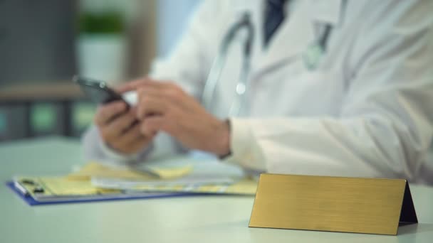 Médico en uniforme enviando mensajes de texto al paciente en el teléfono inteligente, placa de identificación en blanco en la mesa — Vídeos de Stock