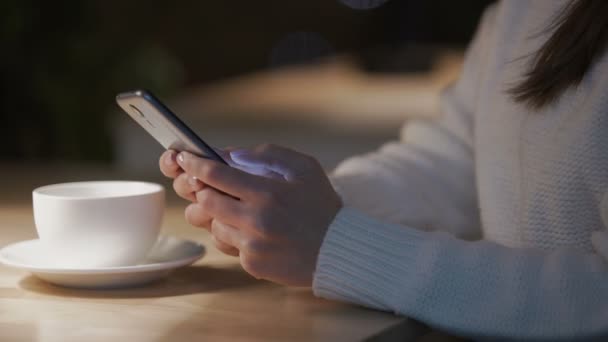 Жінка прокручує сторінки на смартфоні, сидячи в зручному кафе вночі — стокове відео