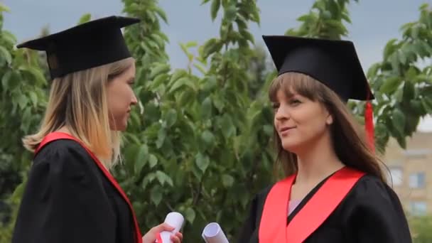 Las mejores amigas en vestidos académicos con diplomas y charlando en el parque — Vídeo de stock