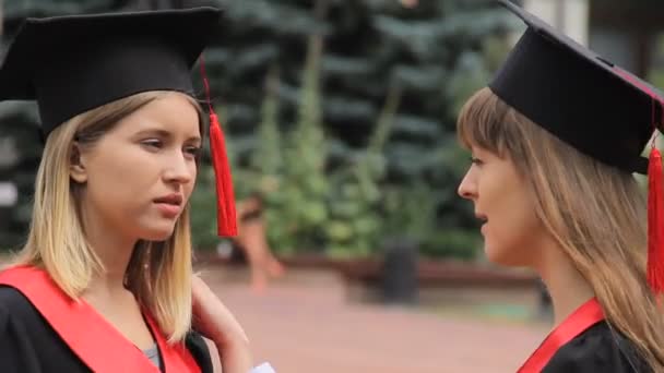 Jovens mulheres bonitas em vestidos acadêmicos conversando perto da universidade, graduação — Vídeo de Stock