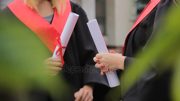 Estudiantes graduadas sosteniendo diplomas atados con cintas rojas y charlando — Vídeo de stock