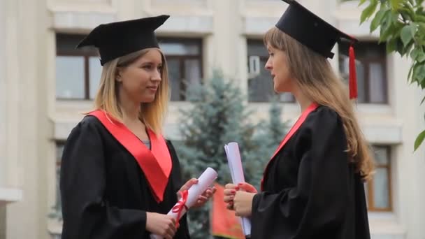 Счастливые улыбающиеся выпускники общаются рядом с академией и держат дипломы, беседуют — стоковое видео