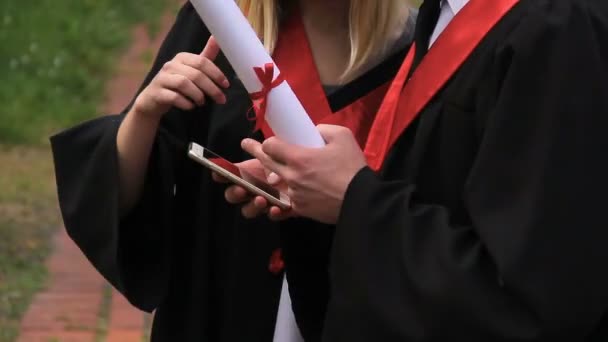 Serious male and female students in academic dresses reading news on smartphone — Stock Video