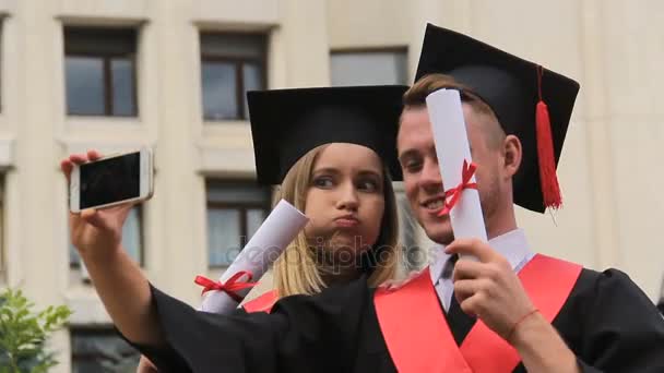 Estudiantes divertidos en vestidos académicos tomando selfies por teléfono, ceremonia de graduación — Vídeos de Stock